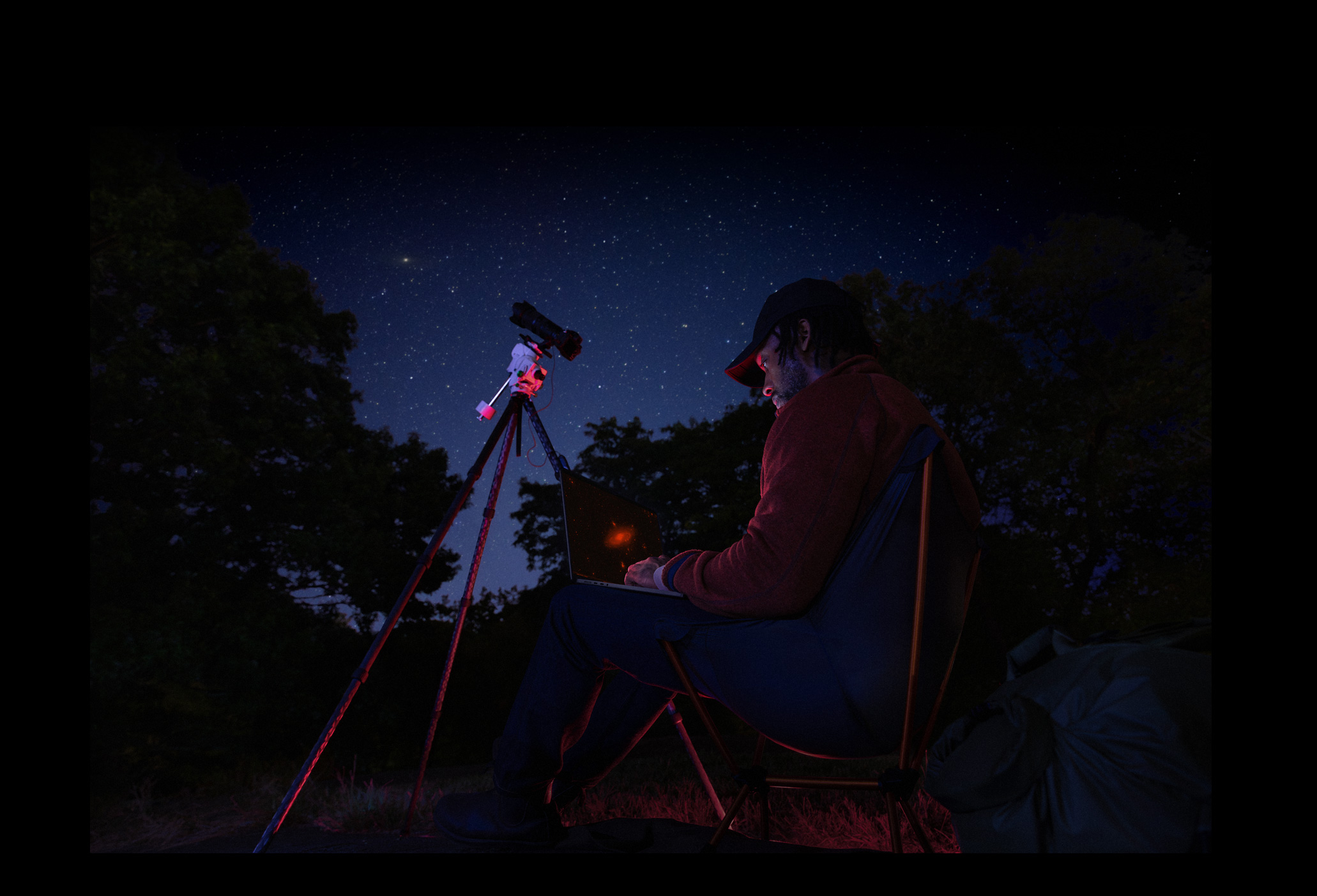 夜晚的户外,一个人将 MacBook Pro 放在腿上使用,MacBook Pro 连接着一部安装在三脚架上对准星空的设备。